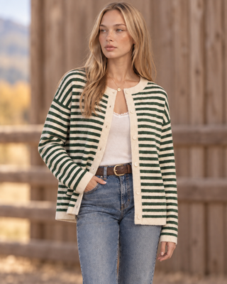 A woman wearing a green and cream striped knit cardigan over a white top and blue jeans, standing outdoors in front of a wooden fence.