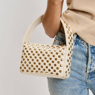 Beige handbag with gold studs held by a person wearing a beige top and blue jeans on a light gray background