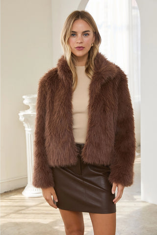 A woman modeling a brown faux fur jacket over a beige top and a dark brown faux leather mini skirt, standing in a bright, modern indoor setting with white columns.