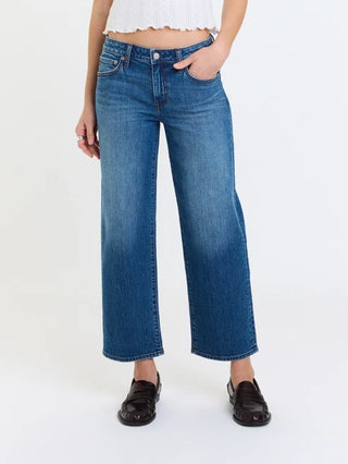 A person wearing medium-wash blue cropped wide-leg jeans with a white cropped top and dark brown loafers, standing against a plain white studio background.