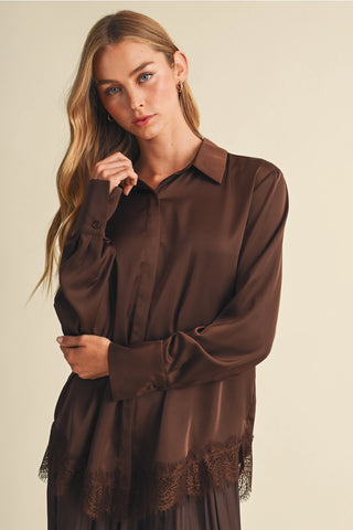 A woman modeling a long-sleeve brown satin blouse with lace trim at the hem, photographed against a plain light beige studio background.
