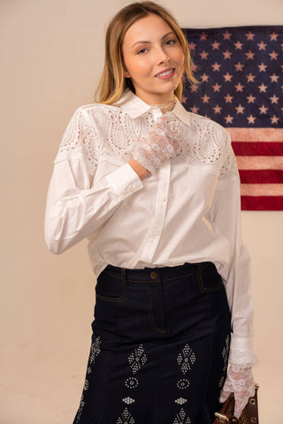 Woman wearing a white blouse with lace details and black pants in front of an American flag.
