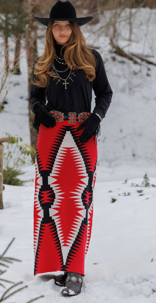 A woman standing outdoors in the snow wearing a long black top, a wide-brim black hat, black gloves, and a long red, black, and white geometric patterned skirt.