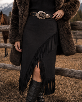 A person wearing a black fringe midi skirt with an asymmetrical hem, styled with a patterned belt, black boots, a brown faux fur coat, and standing in front of a rustic wooden fence with a horse in the background.