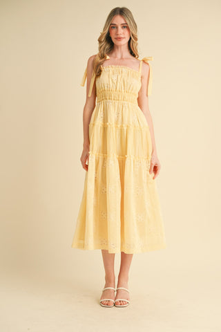 A model wearing a yellow tiered eyelet midi dress with tie strap shoulders, a ruffled neckline, and a flared skirt.