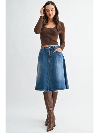 A woman modeling a brown ribbed long-sleeve crop top and a blue denim A-line skirt, paired with brown knee-high leather boots, against a light gray studio background.