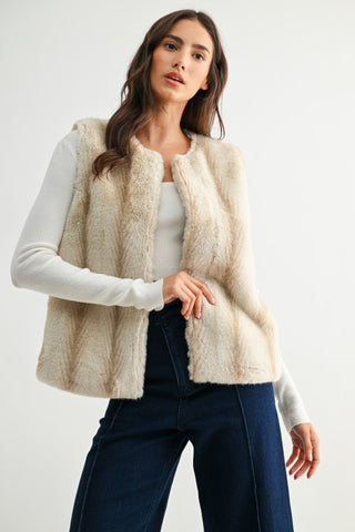 Woman wearing a cream-colored vest over a white long-sleeve shirt and dark jeans on a light gray background