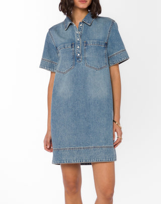 A woman wearing a light blue denim shirt dress with short sleeves, a buttoned collar, and two chest pockets, photographed against a plain white studio background.