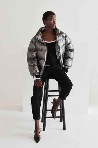 Person wearing a silver puffer jacket sitting on a stool against a white background