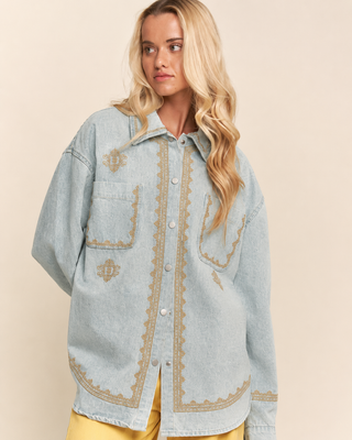 A blonde model wearing a light blue denim button-up shirt with gold embroidered details, paired with yellow pants, against a solid beige background.