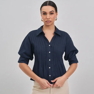 A woman modeling a navy blue pleated button-up blouse with short puff sleeves and white buttons, paired with beige trousers, against a light gray studio background.