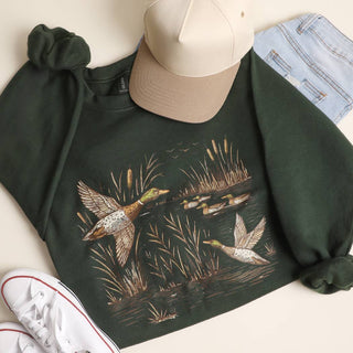 A dark forest green long-sleeve sweatshirt with a detailed graphic of mallards and reeds in a marsh, styled in a flat lay with a beige baseball cap, light blue jeans, white sneakers, and a scrunchie on a light beige background.