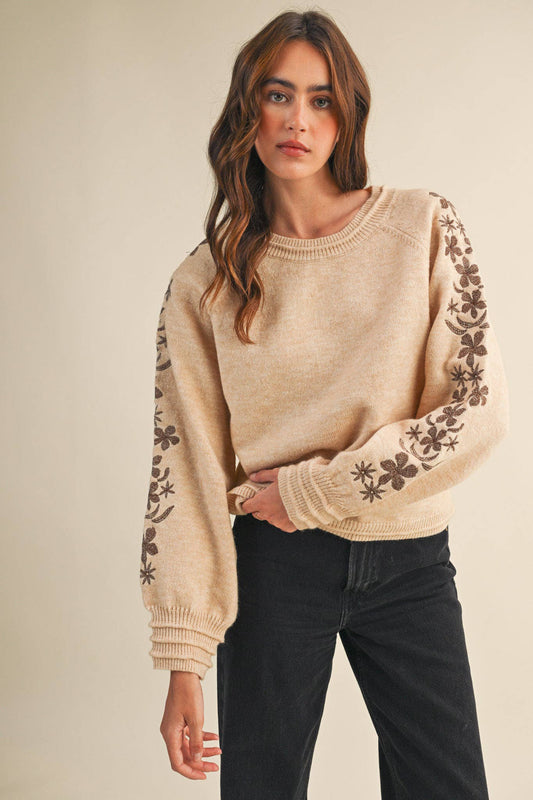 Woman wearing a beige sweater with floral patterns on sleeves against a neutral background