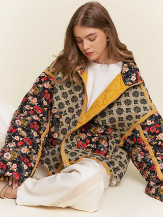 A woman wearing a mixed-media quilted jacket with floral and geometric patterns, mustard yellow trim, and a white top, sitting against a plain cream background.