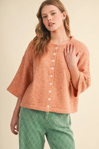 A woman wearing a textured, short-sleeved button-up knit sweater in a peachy orange color, paired with green patterned pants, against a solid beige background.