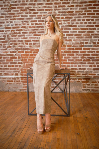 Woman in a beige strapless dress standing on a stool against a brick wall.