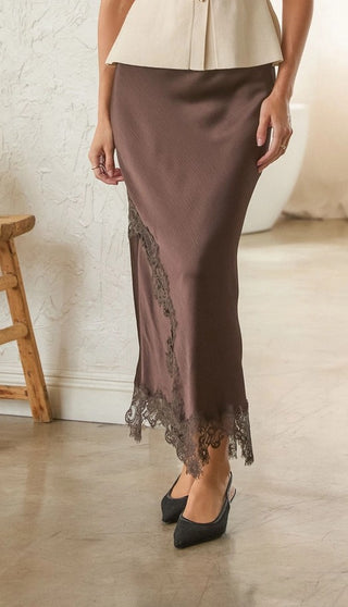 A woman models a brown satin midi skirt with black lace trim along the side and hem, paired with black pointed-toe heels in a bright indoor setting.