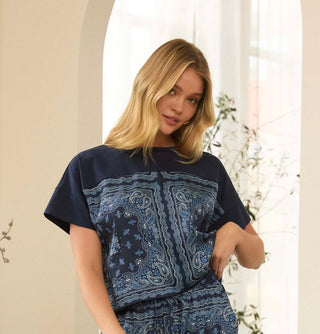A woman wearing a navy blue short-sleeve top with a large paisley bandana print panel across the front, posed in a softly lit indoor setting with a cream-colored wall and window in the background.