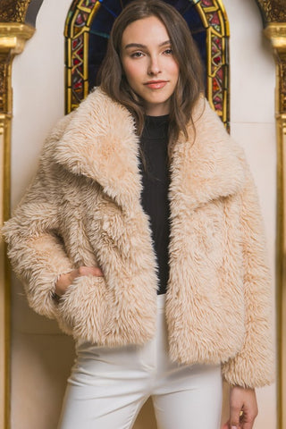 Woman wearing a beige fluffy jacket in front of a decorative wall.