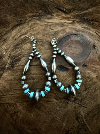 A pair of handmade silver dangle earrings with turquoise and dark metallic beads, displayed on a rustic wooden surface.