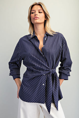 A woman wearing a navy blue and white pinstriped shirt with a tie waist, paired with white trousers, against a light gray studio background.