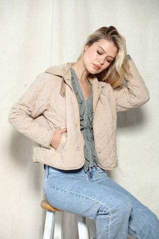 Woman wearing a beige quilted jacket and blue jeans sitting on a stool against a plain background