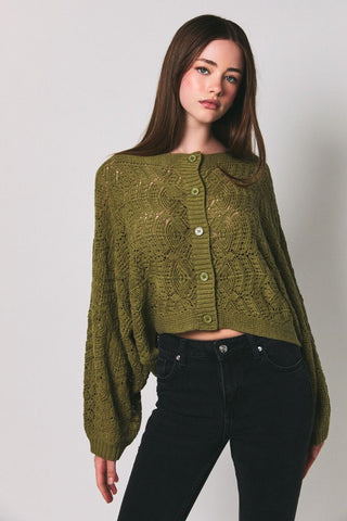 A woman wearing a textured olive green cropped cardigan sweater with balloon sleeves, paired with black pants, against a plain light gray background.
