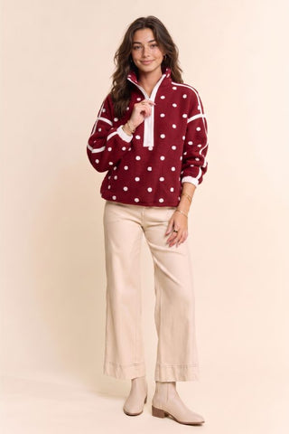 A woman modeling a burgundy and white polka dot quarter-zip sweatshirt with white stripes on the shoulders, paired with beige wide-leg cropped pants and beige ankle boots, against a solid beige studio background.