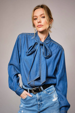 Woman wearing a blue denim shirt with a tie detail and matching jeans against a gray background