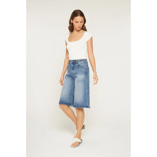 A woman modeling a pair of high-waisted, wide-leg blue denim culottes with a white scoop-neck top, standing against a plain light gray studio background.