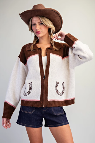 A woman modeling a fuzzy western-style cardigan with brown and cream panels, horseshoe motifs on the front, a brown cowboy hat, and denim shorts against a plain light gray background.