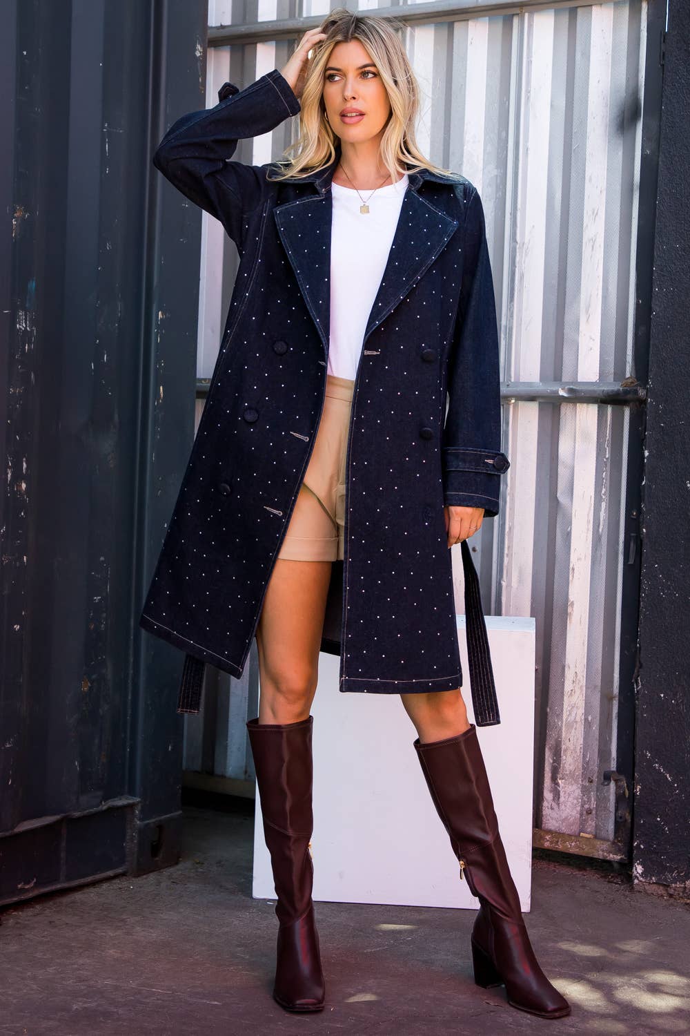 Woman wearing a navy coat with white polka dots, white top, beige shorts, and brown knee-high boots in an urban setting.