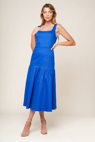 A woman wearing a blue sleeveless, square neckline, ruffled hem midi dress with a back zipper closure.