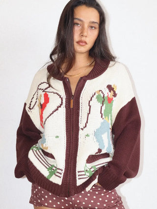 A woman wearing a maroon and cream knit zip-up cardigan with colorful cartoon-style graphics on the front, paired with brown polka-dot shorts, against a plain white background.