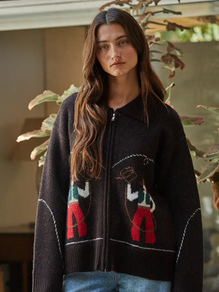 A woman wearing a dark brown zip-up knit cardigan with colorful embroidered figures, standing in front of a softly blurred indoor background with plants.