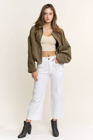 Woman wearing a green jacket, beige top, white pants, and black boots on a plain background