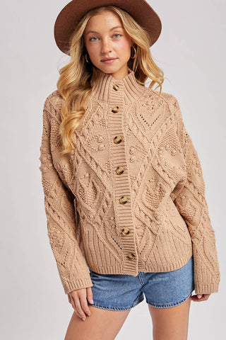 Woman wearing a beige cable knit cardigan, denim shorts, and a brown hat on a light gray background