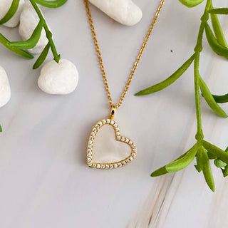 A gold-colored heart-shaped locket necklace with a mother of pearl insert and cubic zirconia accents, displayed on a white surface with green leaves and small white stones in the background.