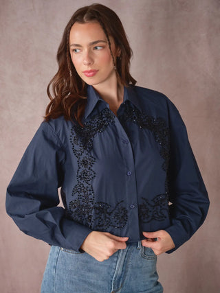 A woman wearing a navy blue long-sleeve button-up shirt with black beaded western-style embroidery on the chest and front, paired with blue jeans, posed against a textured brown studio backdrop.