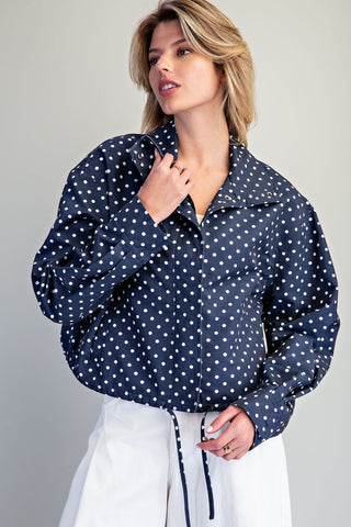 A woman wearing a navy blue long-sleeve button-up shirt with a white polka dot pattern and white high-waisted pants, posed against a neutral light gray background.