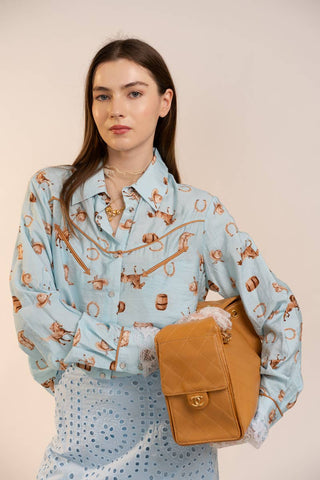 Woman wearing a light blue patterned blouse holding a tan handbag against a beige background