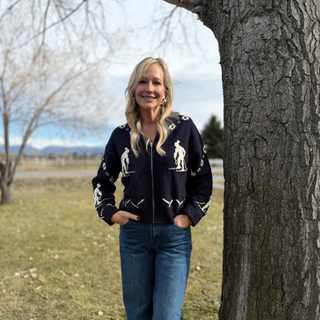Woman wearing a black jacket with white patterns standing next to a tree in an outdoor setting.