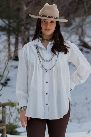 A woman wearing a white long-sleeve button-up blouse with vertical pleats, a beige wide-brim fedora hat, layered silver necklaces, and brown pants, standing outdoors in a snowy setting.