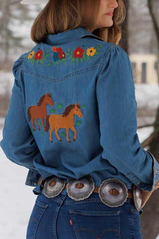 A woman wearing a blue denim western-style jacket with embroidered horses and flowers on the back, shown outdoors in a snowy setting.
