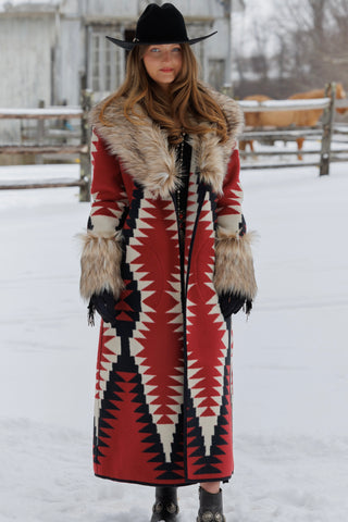 A woman stands outdoors in a snowy setting wearing a long red, black, and cream geometric patterned coat with a large beige fur collar and matching fur-trimmed cuffs, along with a black wide-brim hat.