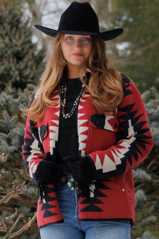 A woman wearing a red, black, and white geometric patterned cardigan, a black cowboy hat, black gloves, and blue jeans, standing outdoors in front of evergreen trees.