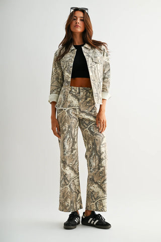 A model stands against a light gray studio background wearing a matching camouflage jacket and wide-leg pants set, styled with a black crop top, black sneakers, and sunglasses on her head.