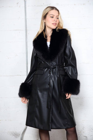 Woman wearing a black coat and black leather shorts against a white brick wall.