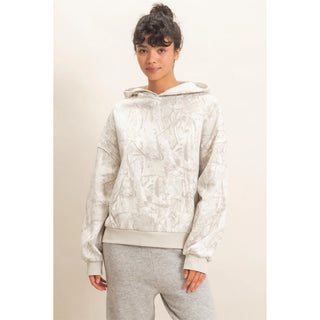 A woman modeling a light beige and white patterned hooded sweatshirt with matching gray sweatpants, set against a plain beige studio background.