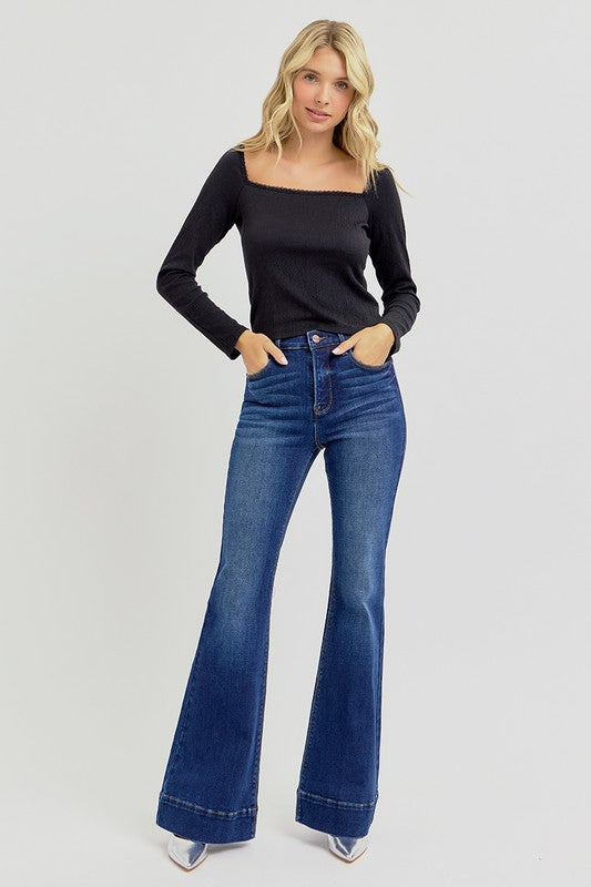Woman wearing a black long-sleeve top and blue flared jeans on a light gray background